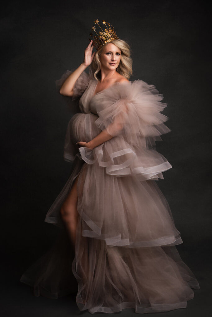 Maternity Photographer, a pregnant woman wears a luxurious dress and holds acrown on her head confidently